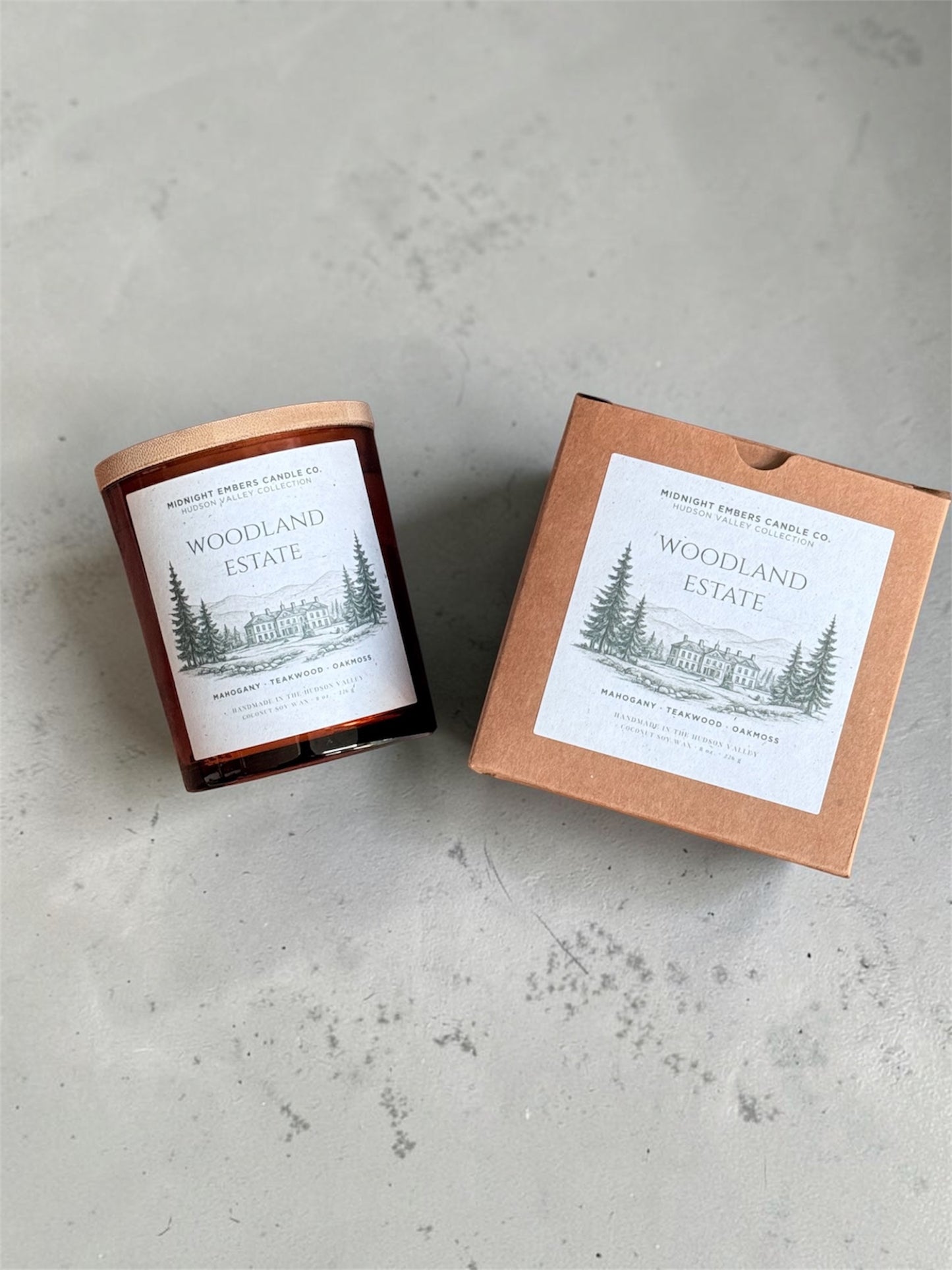A glass jar candle with a copper-toned lid sits beside its kraft paper box on a light gray textured surface. Both pieces display a white label with a black line drawing of pine trees and the text 'Woodland Estate' and 'Midnight Forest Candle Co.