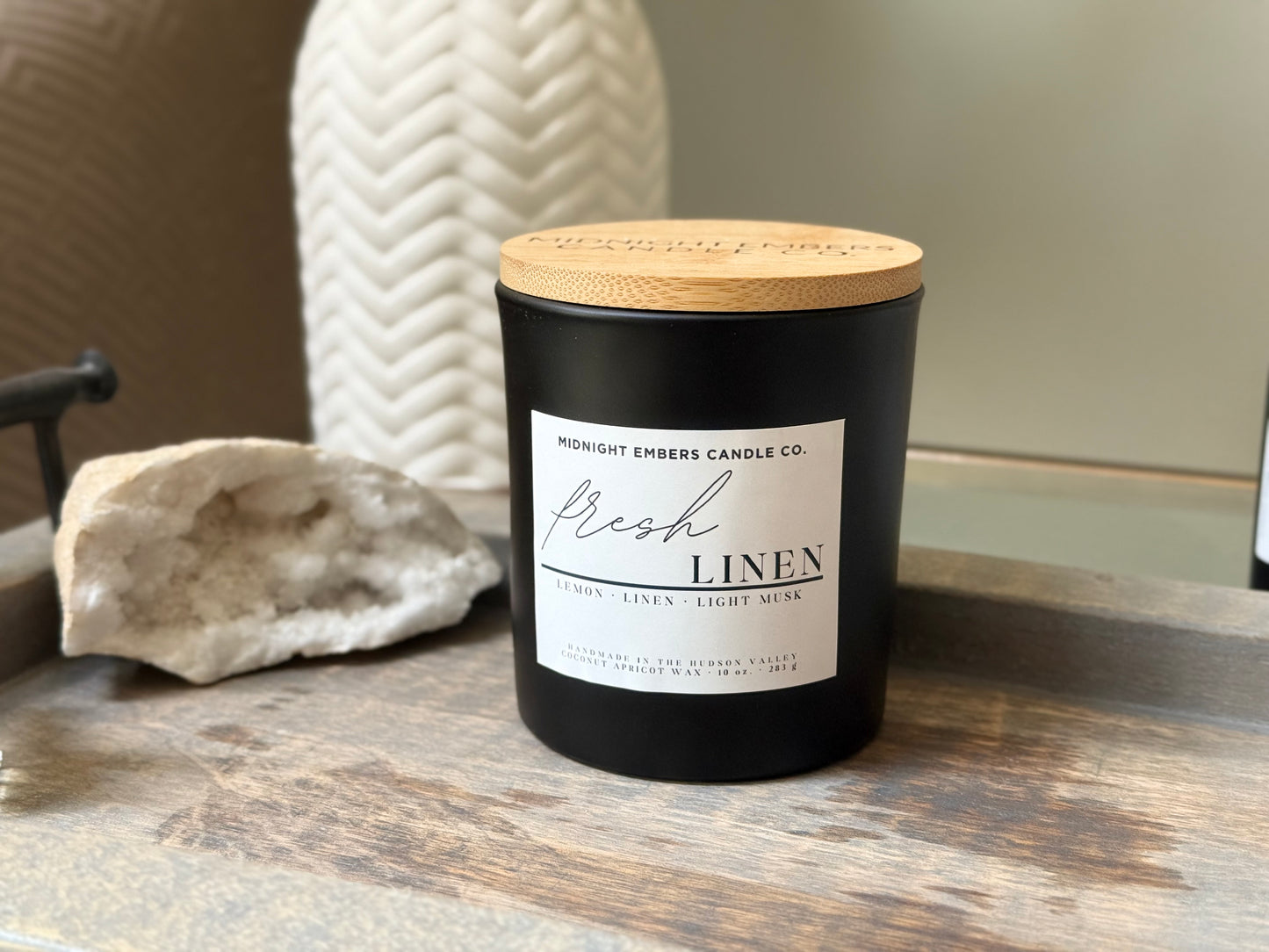 A black candle with a bamboo wooden lid labeled 'Fresh Linen', set on a wooden surface with a white textured object to the side.
