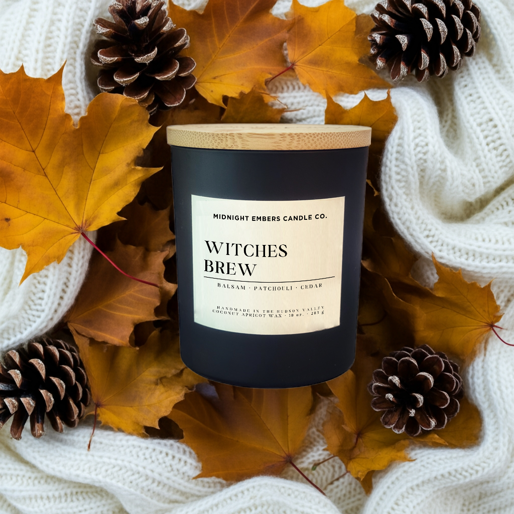 Candle labeled 'Witches Brew' surrounded by autumn leaves and pinecones on a white textured fabric background.