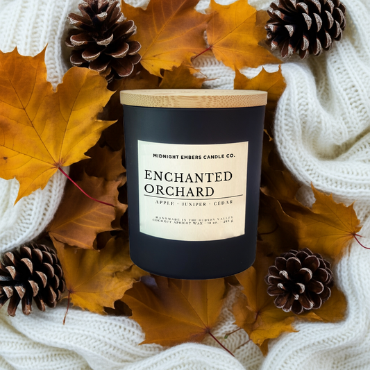 Candle labeled 'Enchanted Orchard' surrounded by autumn leaves and pinecones on a white textured fabric background.