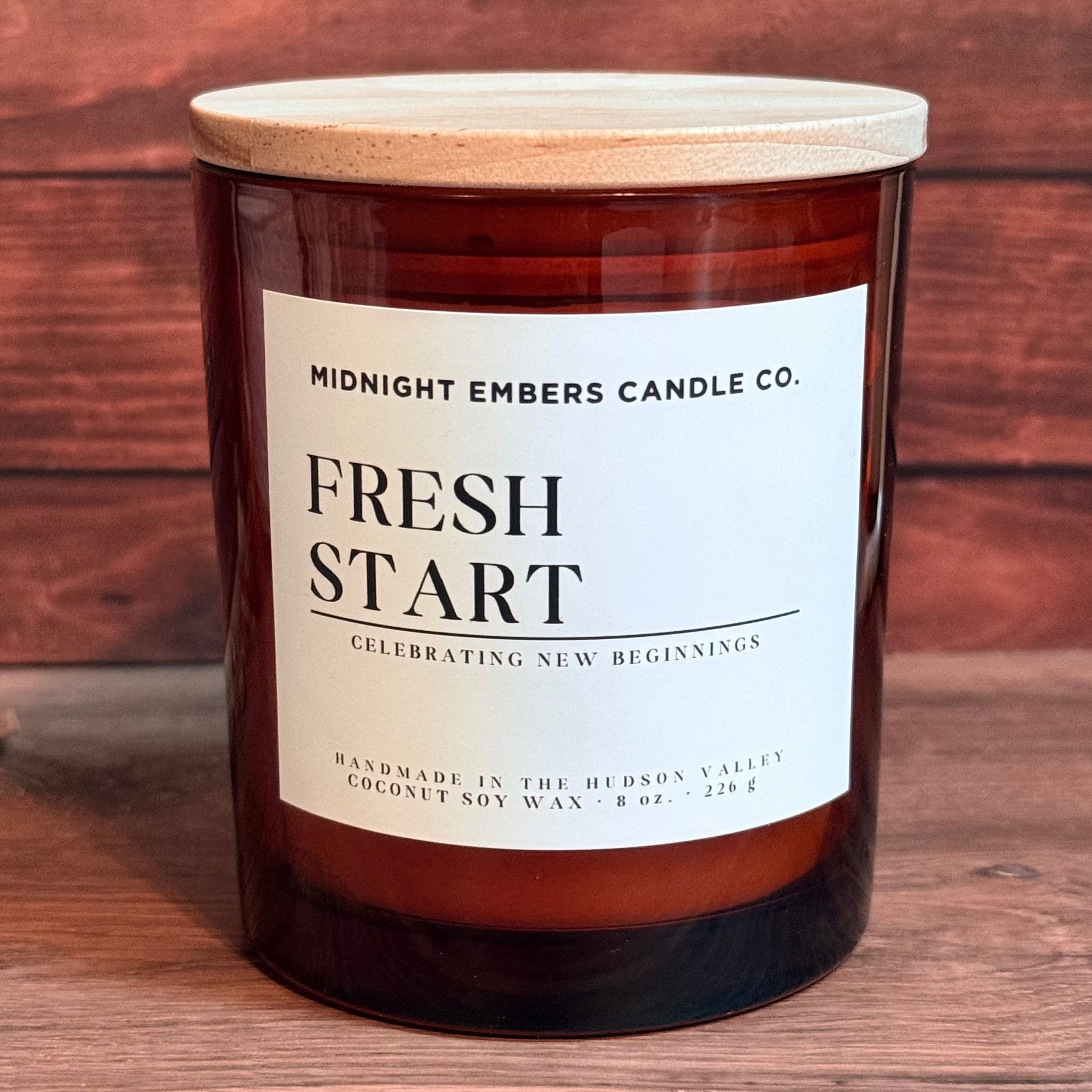 A jar candle with a light wood lid and a white label reading 'FRESH START' by Midnight Embers Candle Co., set on a wooden surface against a rustic wood plank background.