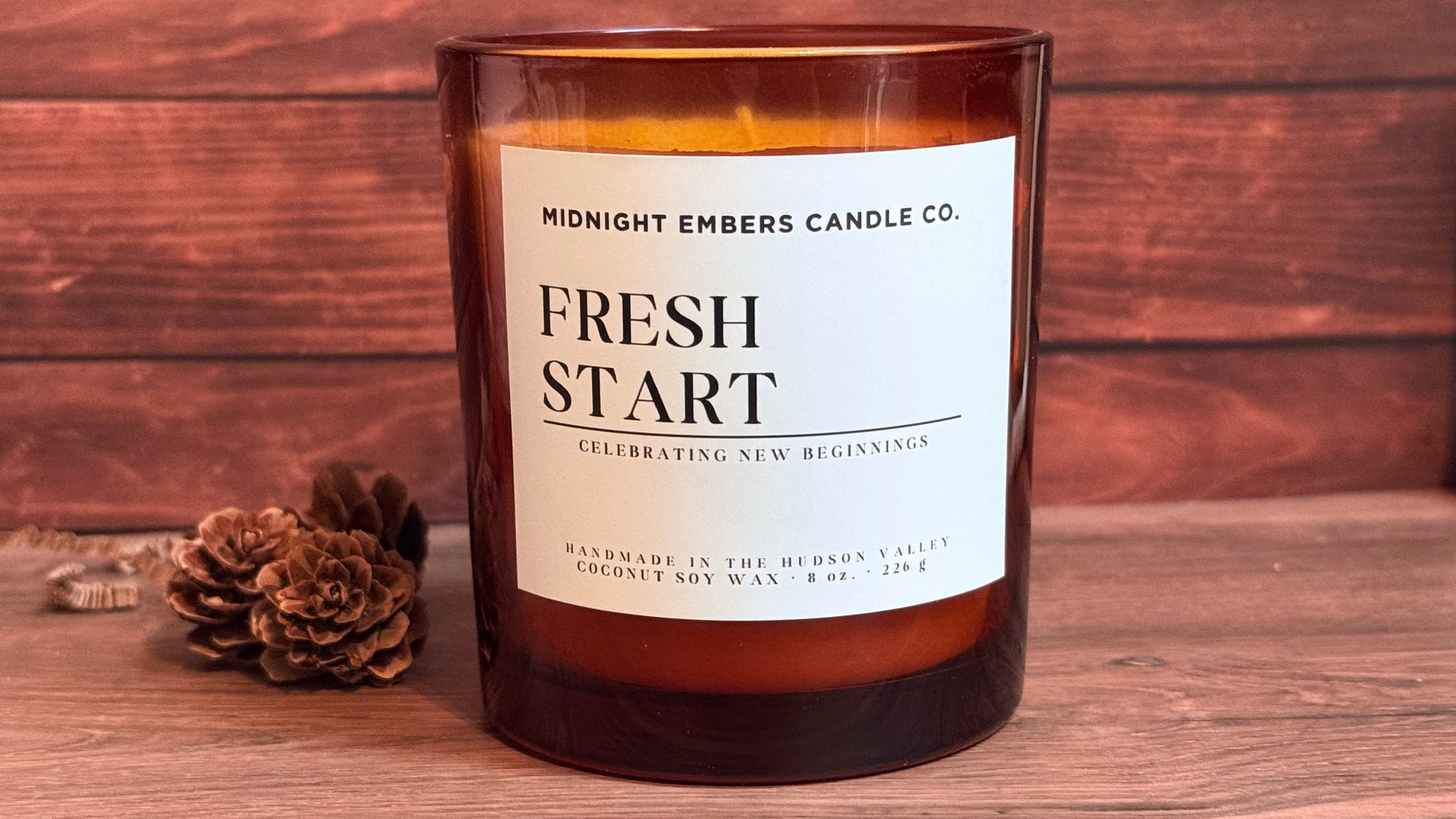 A candle in a dark amber glass jar with a white label reading 'MIDNIGHT EMBERS CANDLE CO. FRESH START' on a wooden surface, with pinecones beside it and a rustic wood plank background.