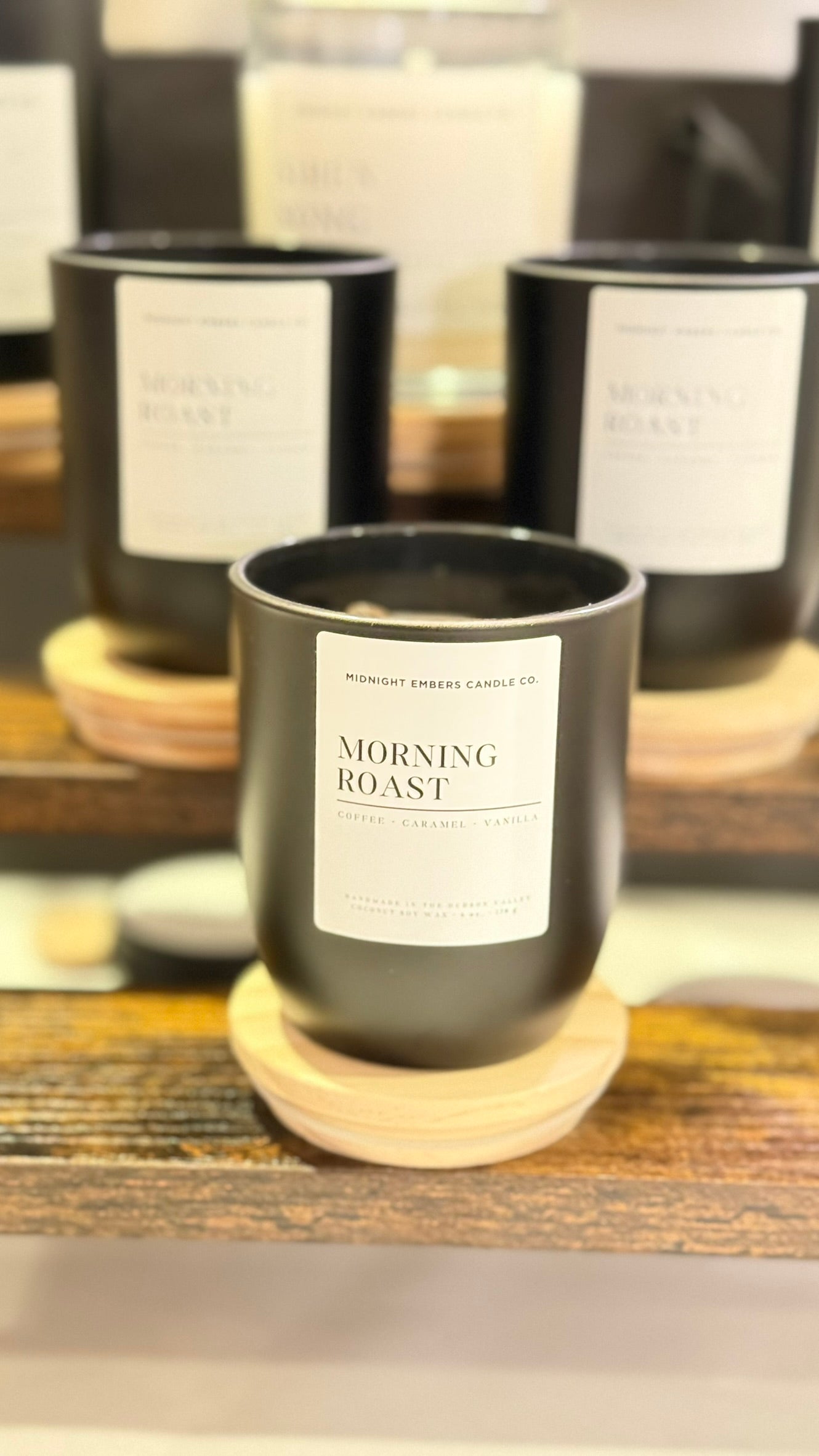 Candle labeled 'Morning Roast' on a wooden coaster with more candles in the background.