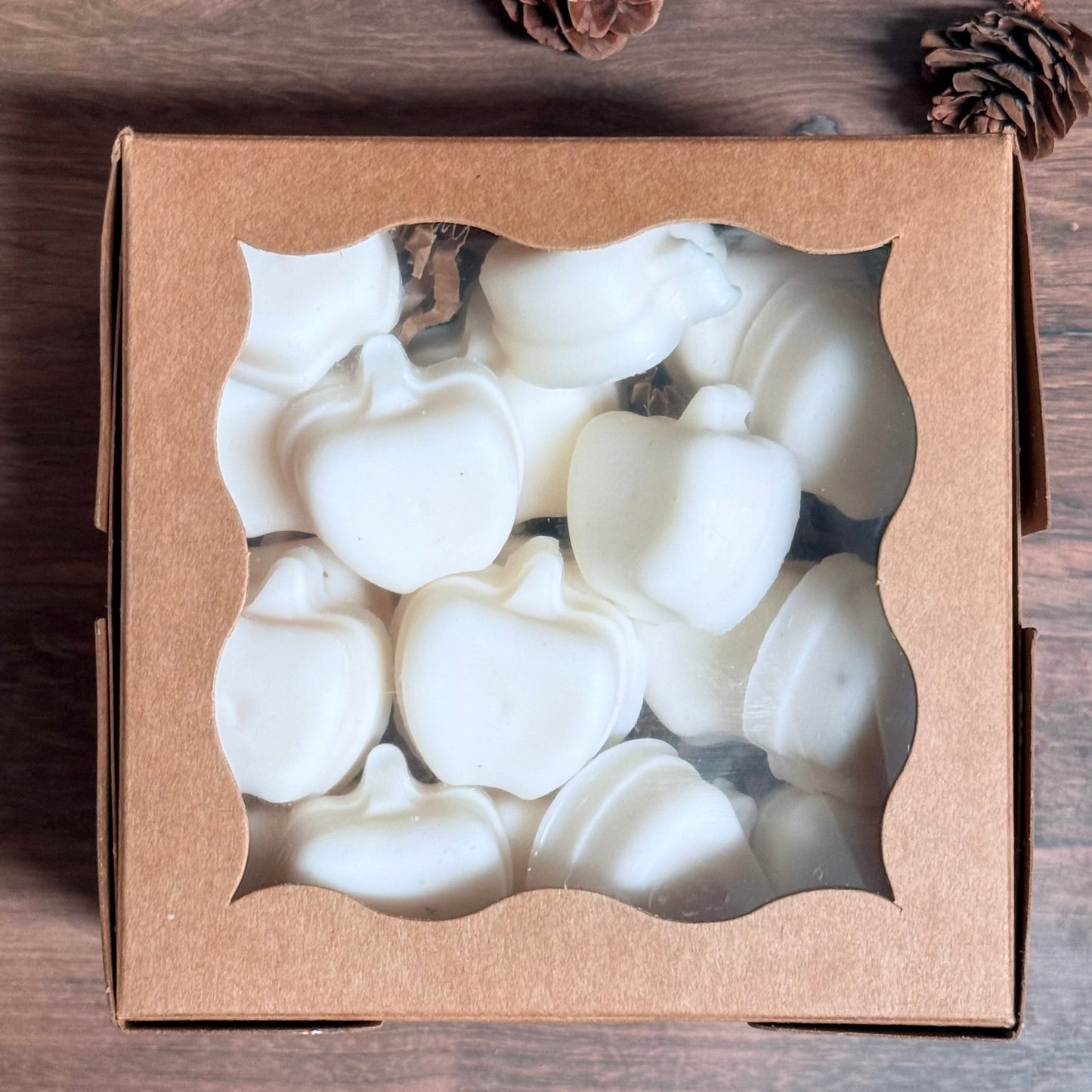 A square brown kraft gift box with a clear window showing white pumpkin-shaped wax melts or soap pieces inside, placed on a dark wood surface with pinecones nearby.