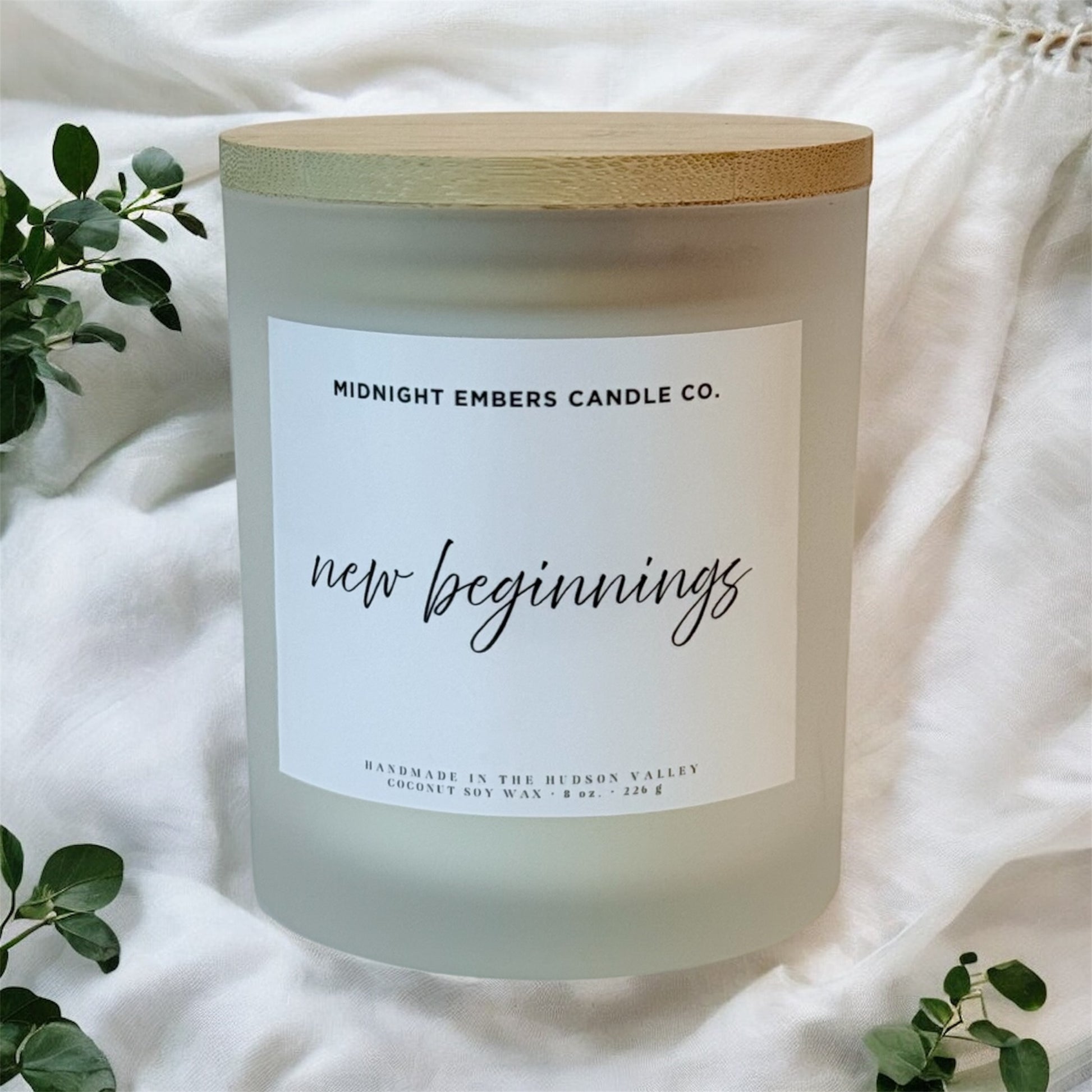 Frosted candle with wooden lid labeled 'Midnight Embers Candle Co. new beginnings' on a white fabric background.