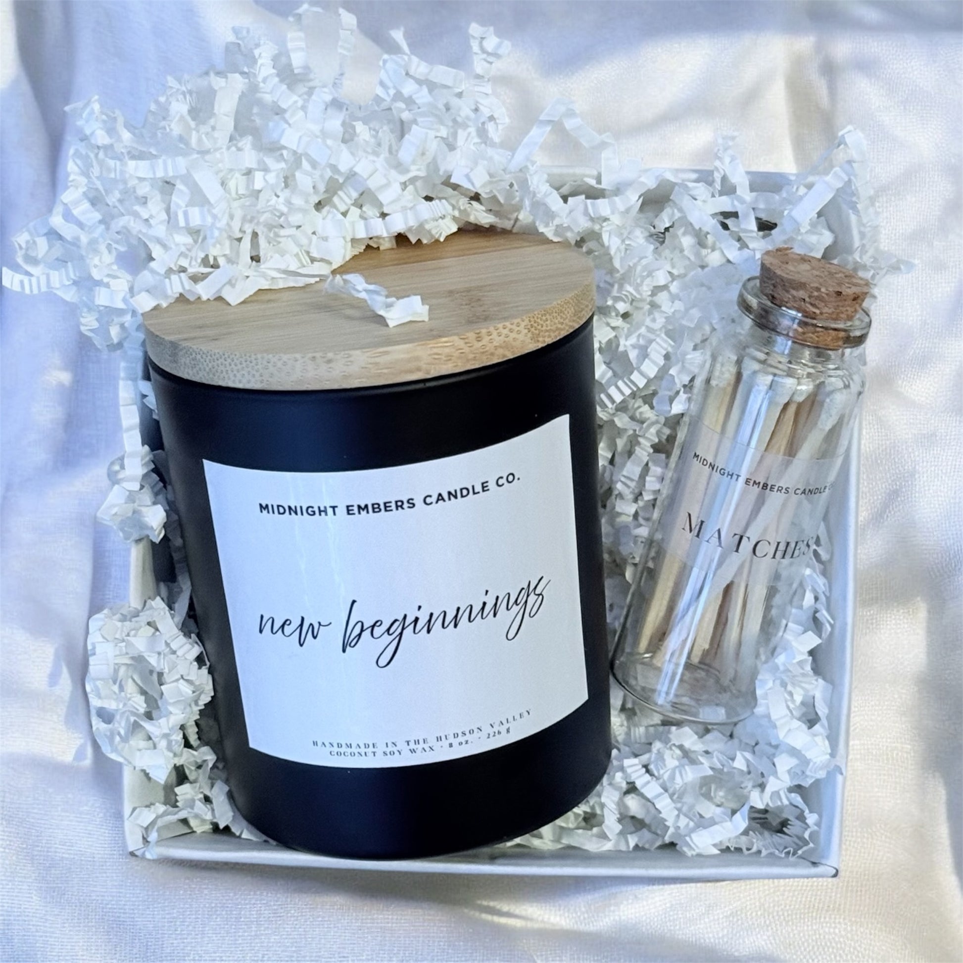 Candle with a wooden lid and match container labeled 'Midnight Embers Candle Co.' on crinkled paper.