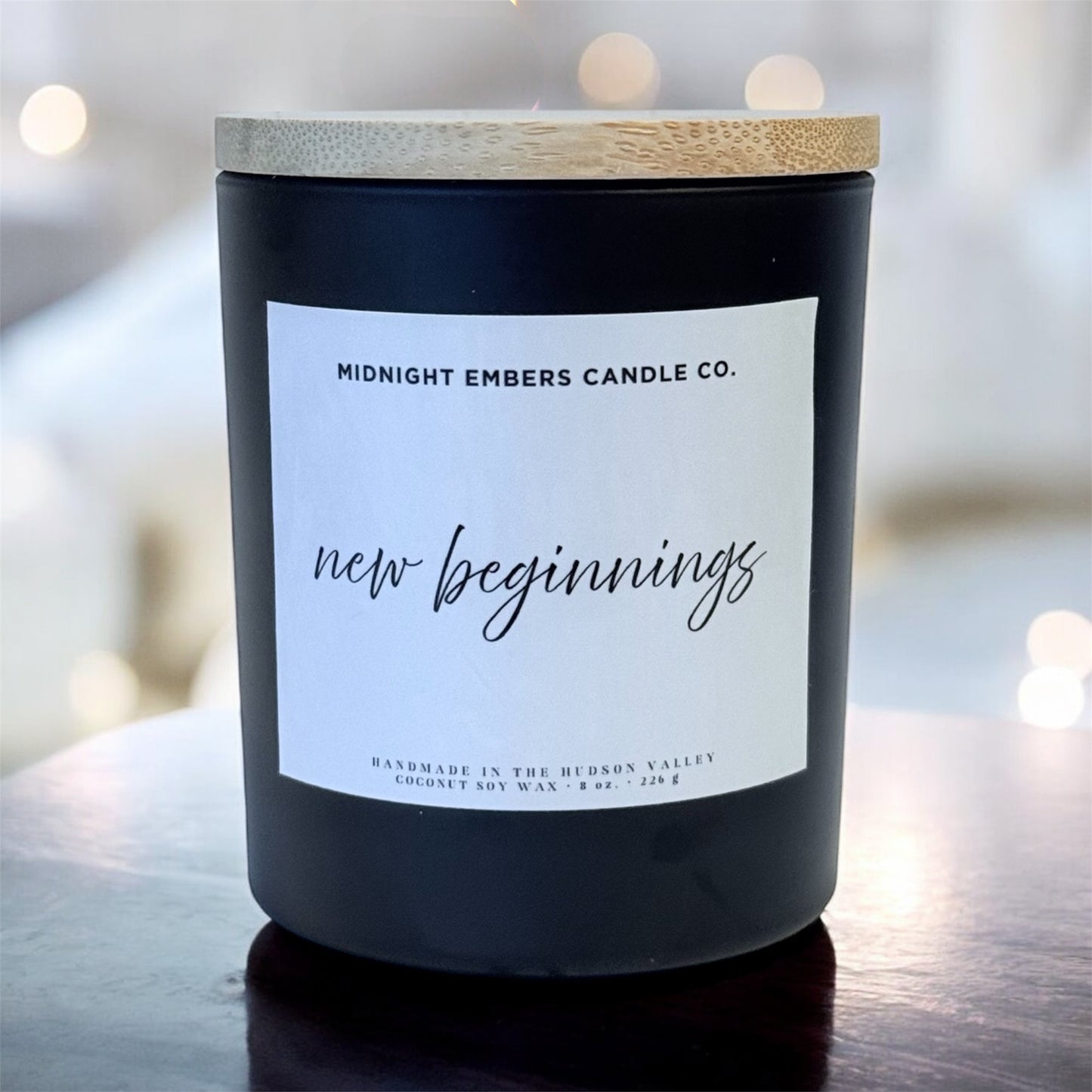 Candle with 'New Beginnings' label on a wooden surface with blurred lights in the background