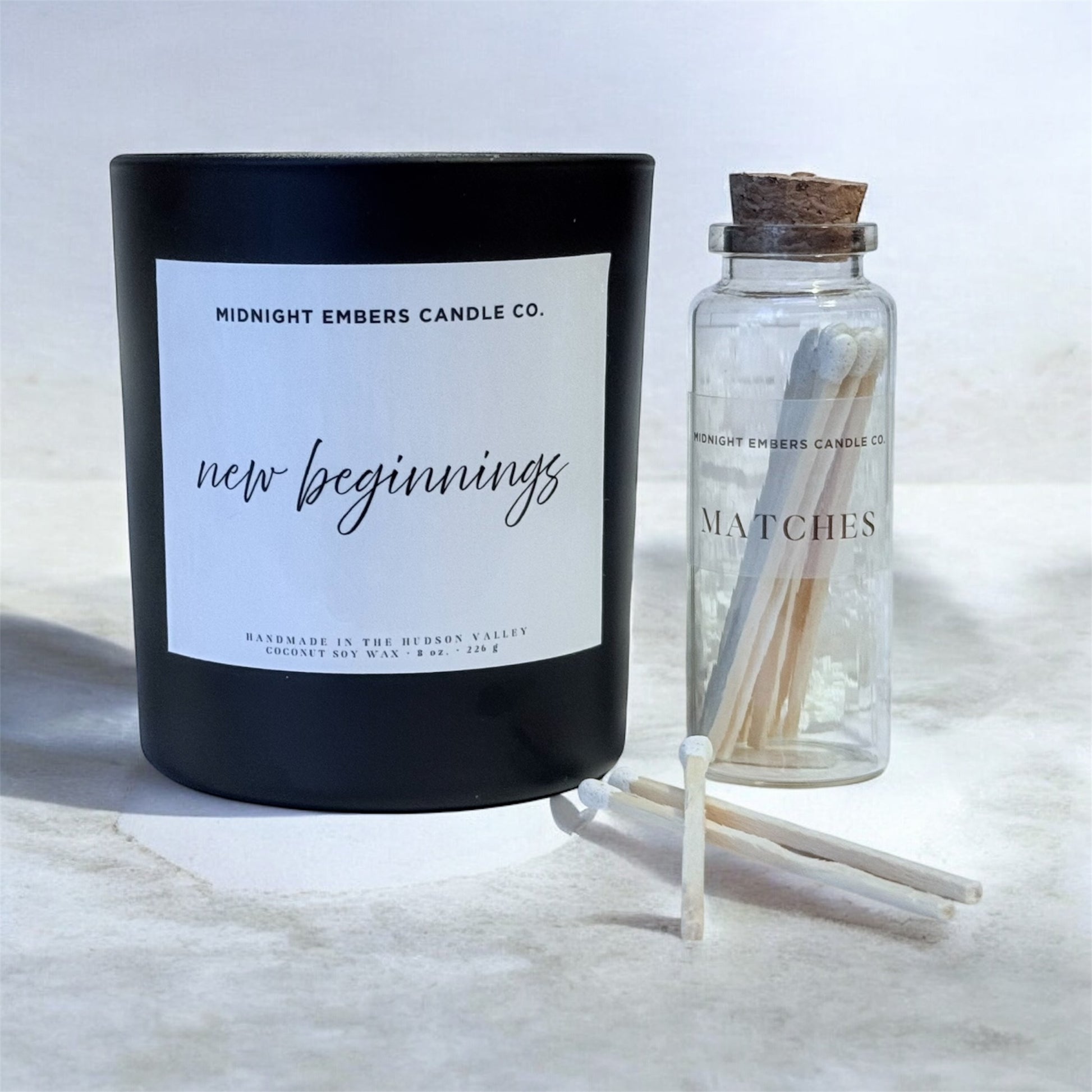 Black candle labeled 'New Beginnings' from Midnight Embers Candle Co. with a bottle of matches on a light background.