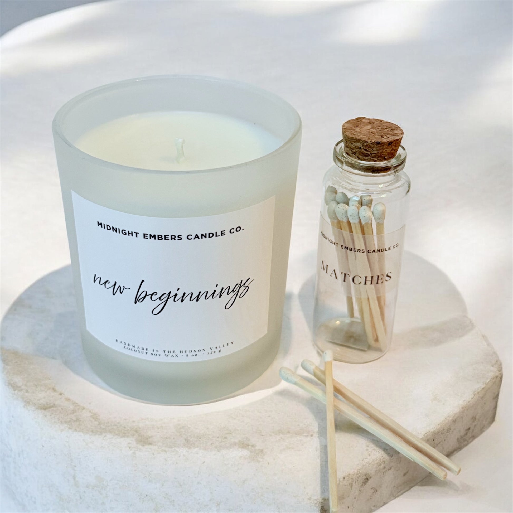 Candle labeled 'new beginnings' with a bottle of matches on a white surface