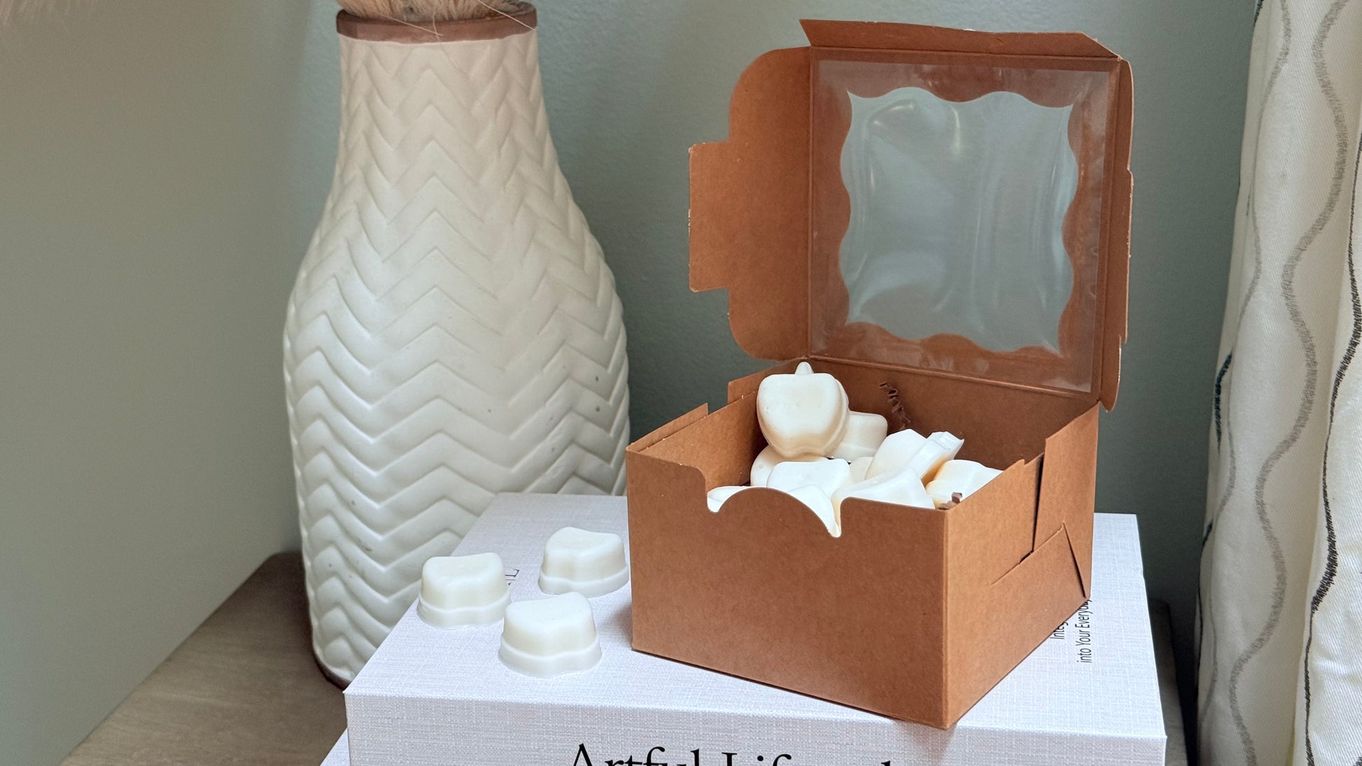 A brown kraft paper gift box with a clear window lid, filled with white ceramic soap bars, displayed on a white book next to a textured white ceramic vase.