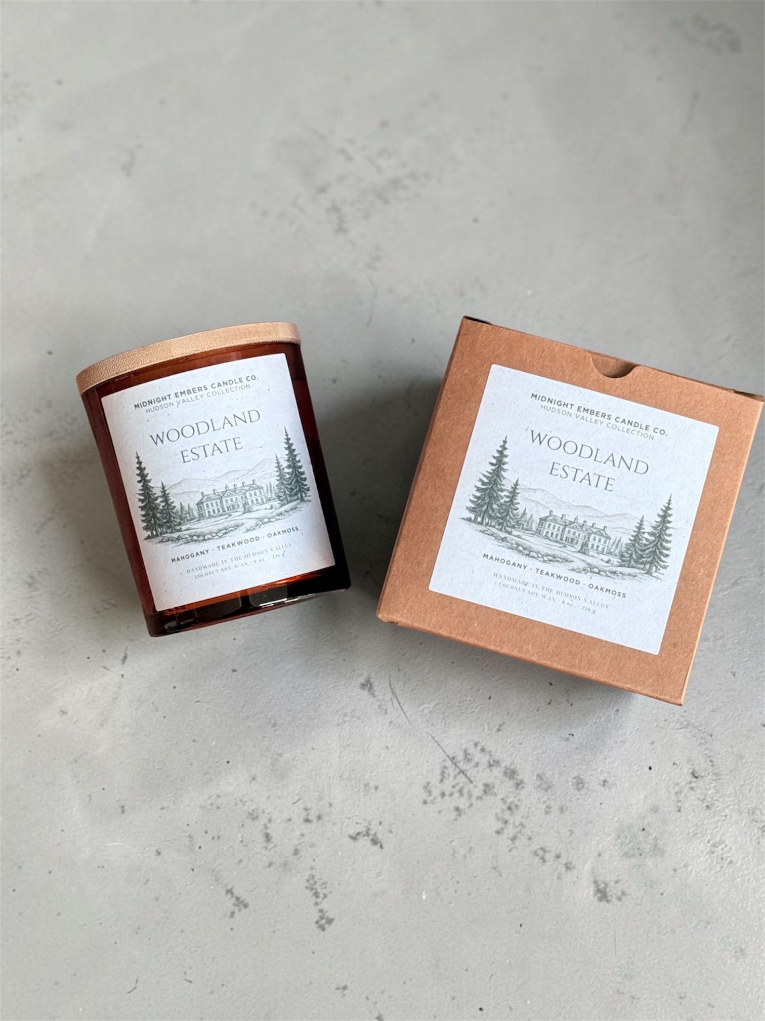 A glass jar candle with a copper-toned lid sits beside its kraft paper box on a light gray textured surface. Both pieces display a white label with a black line drawing of pine trees and the text 'Woodland Estate' and 'Midnight Forest Candle Co.