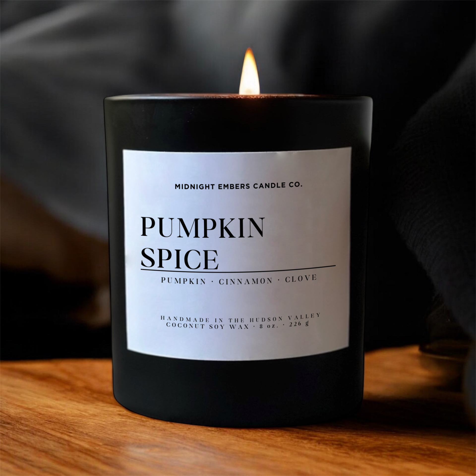 A lit candle in a matte black jar with a white label reading 'PUMPKIN SPICE' and 'MIDNIGHT EMBERS CANDLE CO.' on a wooden surface against a dark fabric background.