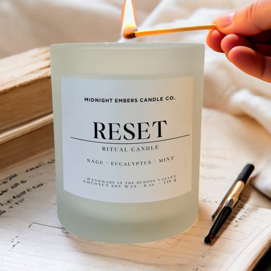 Candle labeled 'Reset' from Midnight Embers Candle Co. being lit with a match on a wooden surface.