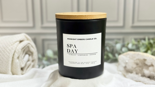 Black candle labeled 'Spa Day' with a wooden lid on a white surface, with a blurred background of greenery and towels.