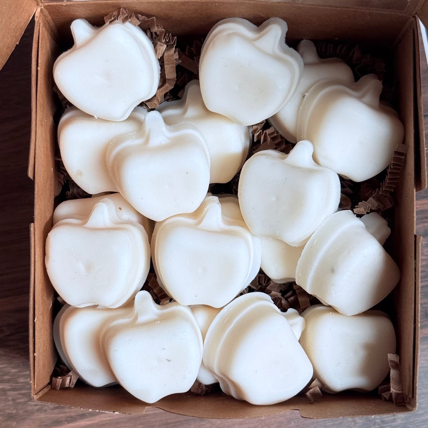 White apple-shaped wax melts arranged in a brown cardboard gift box with a clear lid, set on a dark wooden surface.