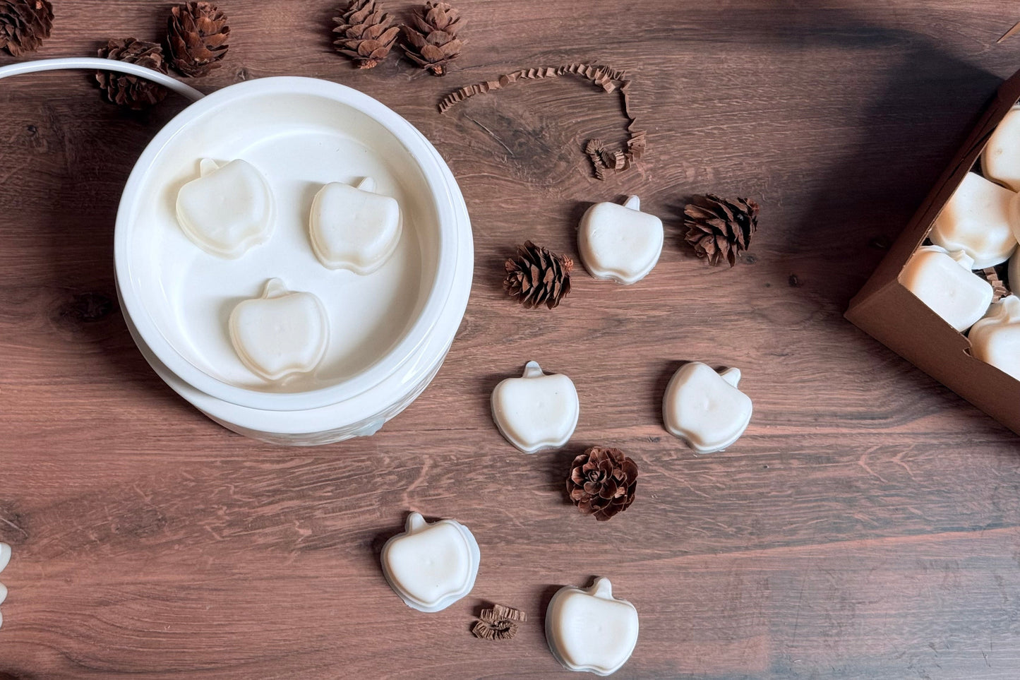 White apple-shaped wax melts displayed in a white round wax melt warmer on a rustic wooden surface, surrounded by pinecones, and a box of additional wax melts.