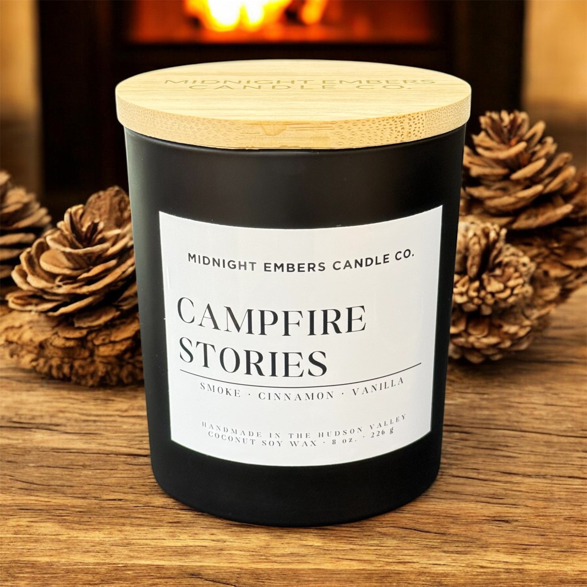 Candle labeled 'Campfire Stories' from Midnight Embers Candle Co. on a wooden surface with pine cones.