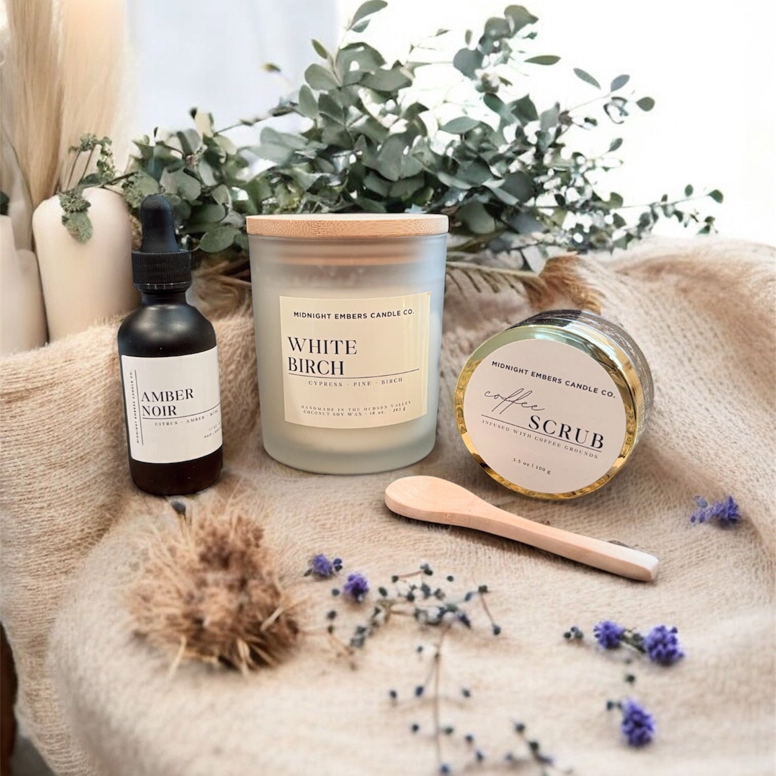 Skincare products including a candle, serum, scrub, and brush on a textured surface with eucalyptus leaves.