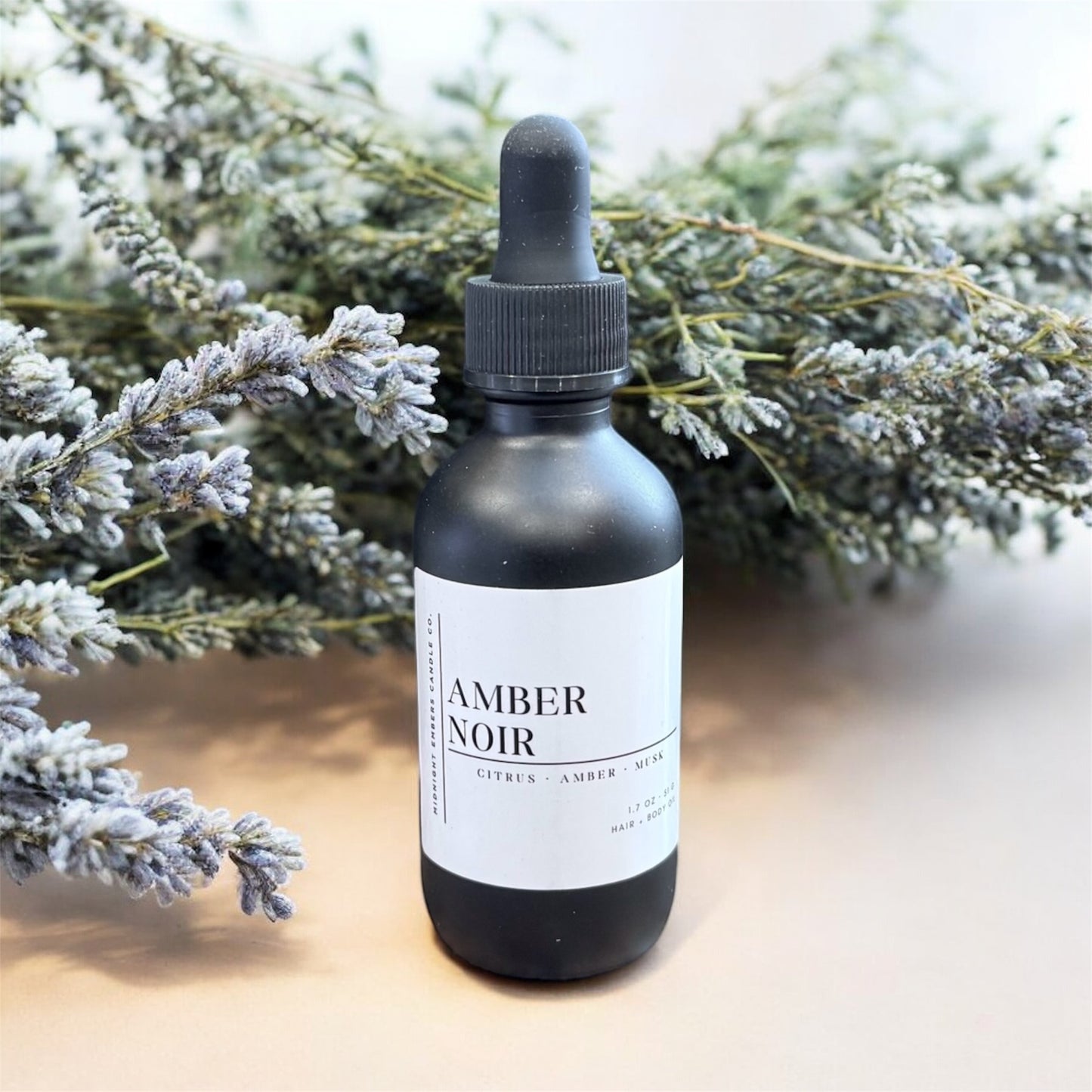 Black bottle labeled 'Amber Noir' with a dropper on a light surface with lavender in the background