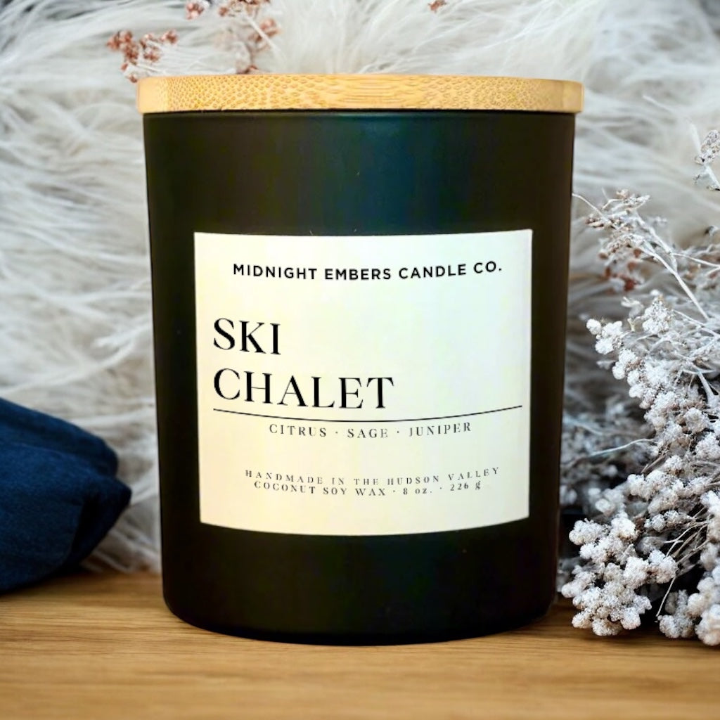 Candle with 'Ski Chalet' label by Midnight Embers Candle Co. on a wooden surface with decorative elements.